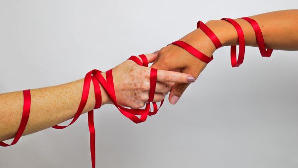 Mulheres segurando a mão uma da outra com fitas vermelhas entrelaçadas representando a conscientização de pessoas vivendo com HIV / Aids