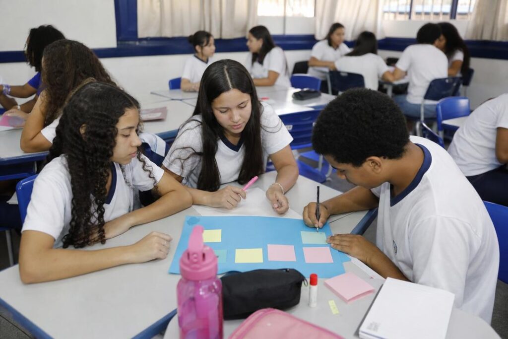 Estudantes uniformizados trabalham em grupo com cartolina e post-its em sala de aula, em atividade de educação empreendedora