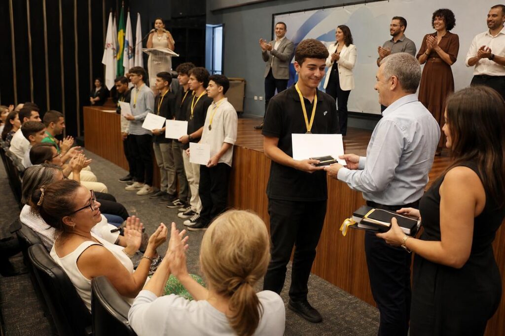 Estudante com medalha recebe certificado das mãos de um representante durante cerimônia de premiação, com colegas premiados e plateia aplaudindo