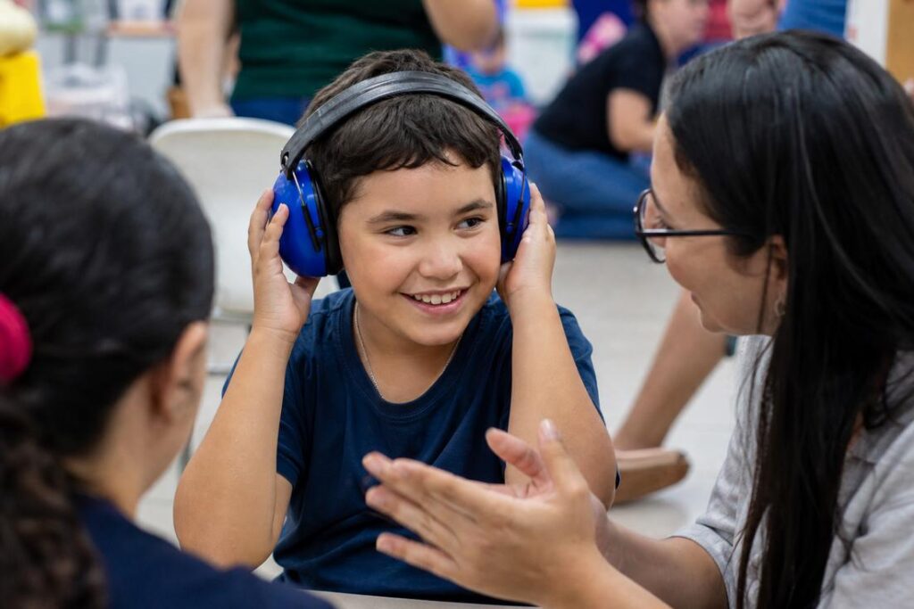 Menino sorrindo usa abafadores de ruído azuis enquanto uma mulher ajusta o protetor auricular, em ambiente interno