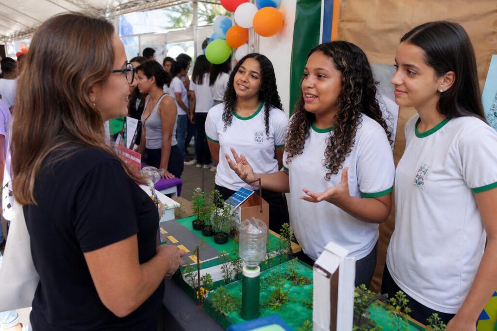 Três estudantes de uniforme apresentam uma maquete de projeto a uma visitante em estande de feira, com balões coloridos ao fundo