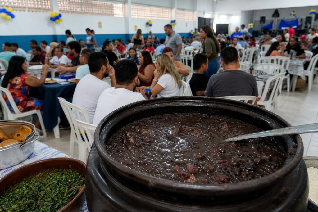 Panela de barro com feijoada em primeiro plano e salão cheio de pessoas ao fundo durante a feijoada da APAE de Timóteo