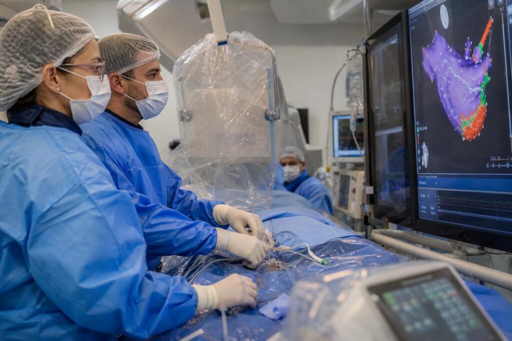 Dois médicos de aventais azuis conduzem a ablação de fibrilação atrial observando o mapa eletroanatômico 3D do coração no monitor, no Hospital Márcio Cunha